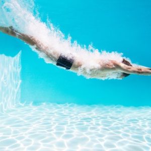 A swimmer in black trunks dives underwater through a bright blue swimming pool, leaving a trail of white bubbles behind.
