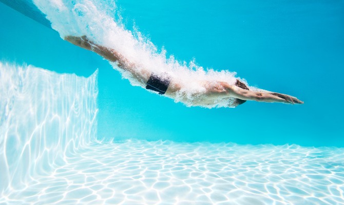 A swimmer in black trunks dives underwater through a bright blue swimming pool, leaving a trail of white bubbles behind.