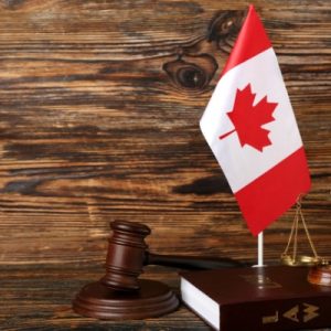 Canadian flag beside a wooden judge's gavel and gold scales of justice resting on a law book against a rustic wood backdrop.