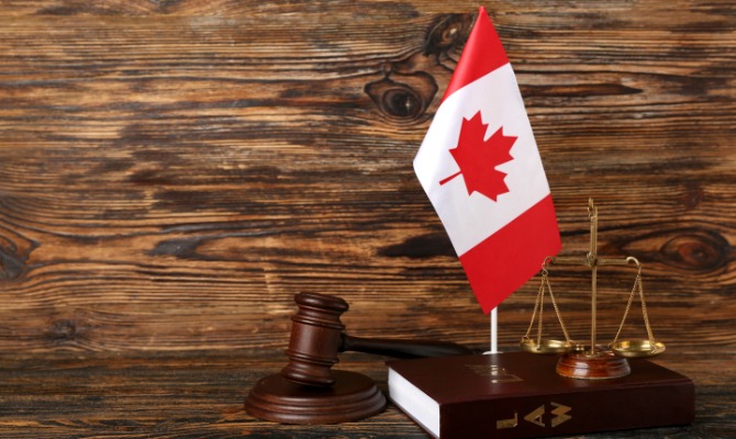 Canadian flag beside a wooden judge's gavel and gold scales of justice resting on a law book against a rustic wood backdrop.