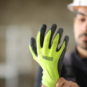 Construction worker wearing a white hard hat adjusts a bright yellow work glove with black rubber-coated fingertips.