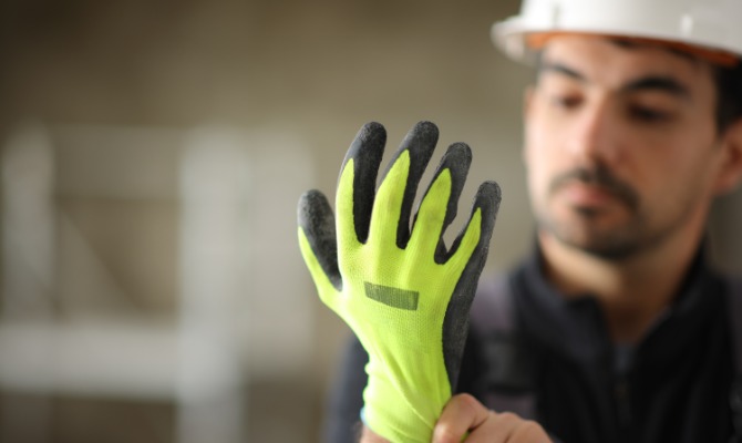 Construction worker wearing a white hard hat adjusts a bright yellow work glove with black rubber-coated fingertips.