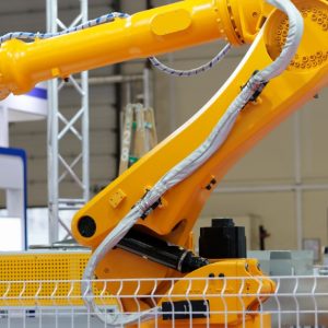 Large yellow industrial robotic arm with a mechanical gripper positioned behind a safety fence in a factory.