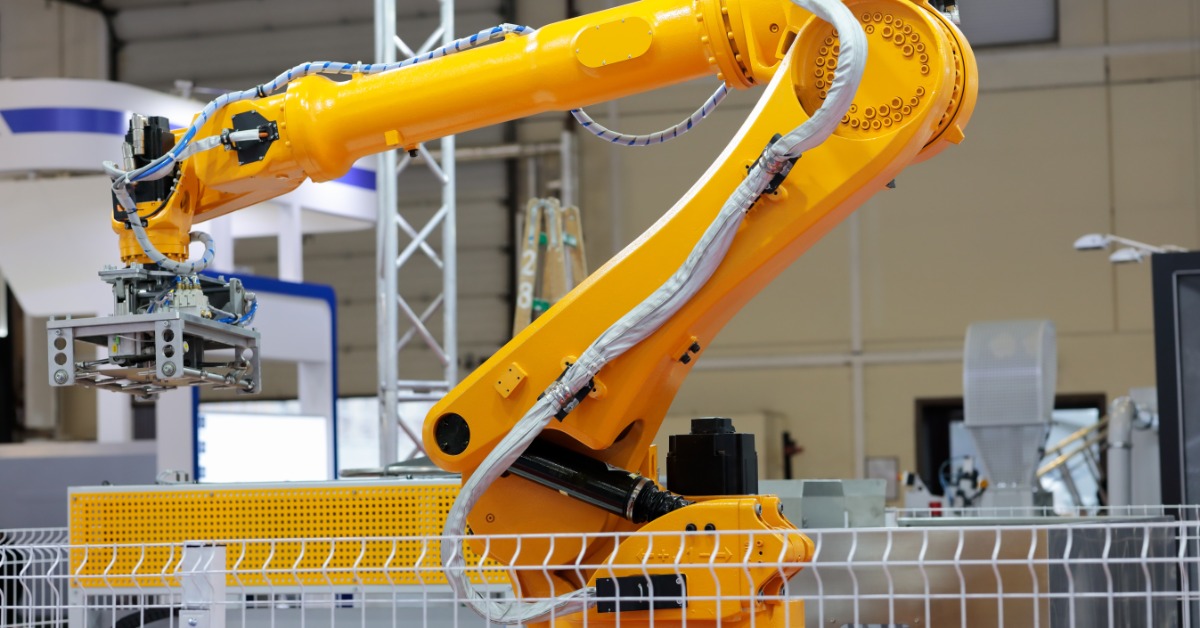 Large yellow industrial robotic arm with a mechanical gripper positioned behind a safety fence in a factory.