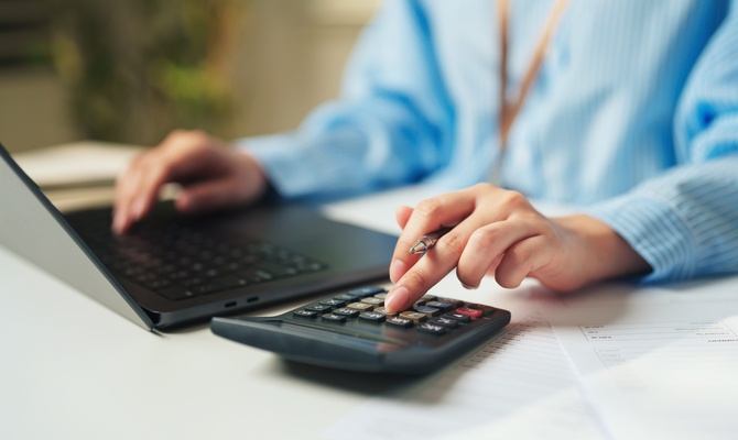 A person in a blue shirt using a calculator and laptop while holding a pen over financial documents on a desk.