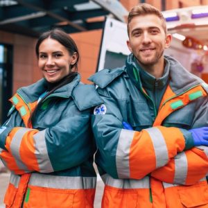 A smiling man and a smiling woman pose with their arms crossed and wearing heavy jackets in front of an ambulance.