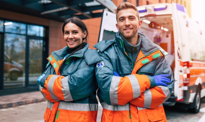 A smiling man and a smiling woman pose with their arms crossed and wearing heavy jackets in front of an ambulance.