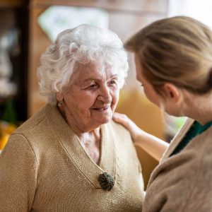 A senior woman wearing a brown sweater standing in front of a woman with her arm holding her in a living room.