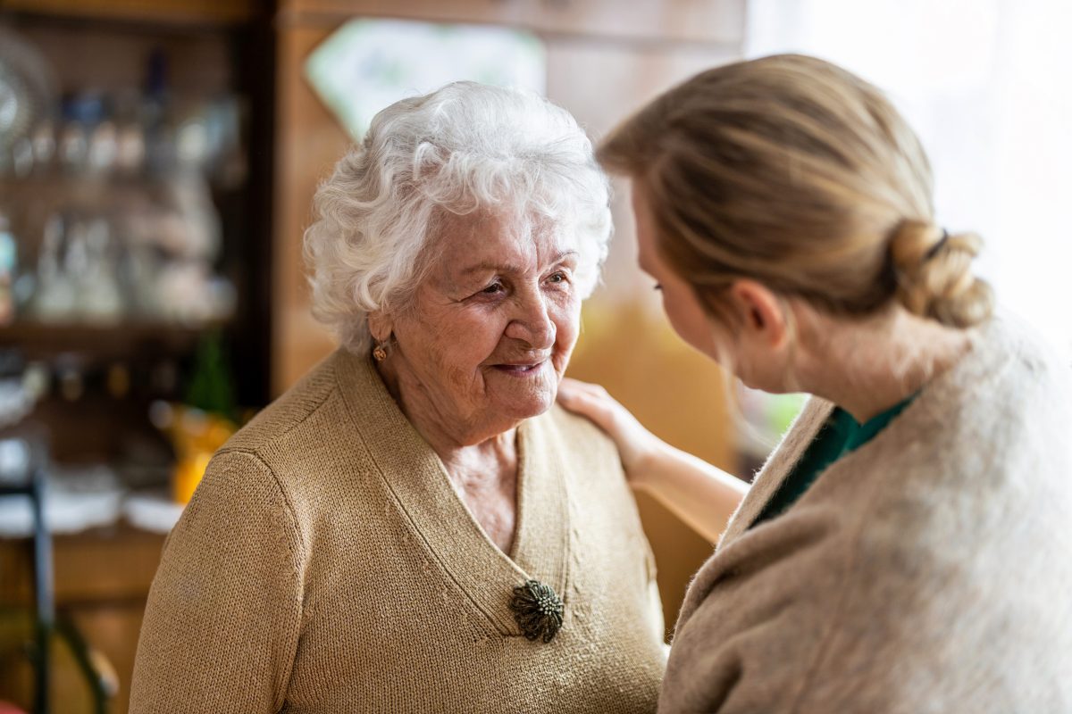 A senior woman wearing a brown sweater standing in front of a woman with her arm holding her in a living room.